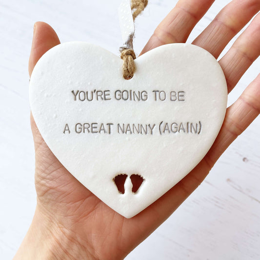 Pearlised white clay hanging heart with baby feet cut out of the bottom and grey personalisation, the heart is personalised with YOU’RE GOING TO BE GREAT NANNY (AGAIN)