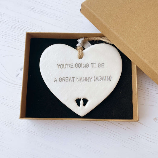 Pearlised white clay hanging heart with baby feet cut out of the bottom and grey personalisation, the heart is personalised with YOU’RE GOING TO BE GREAT NANNY (AGAIN)