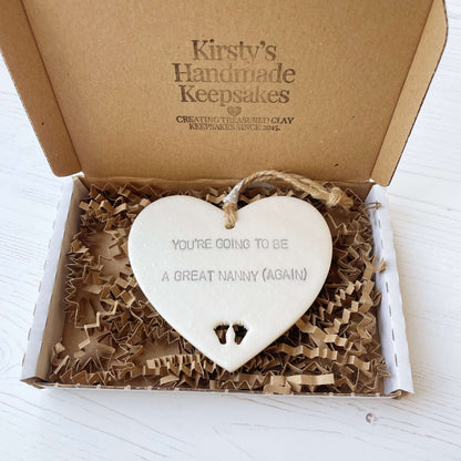 Pearlised white clay hanging heart with baby feet cut out of the bottom and grey personalisation, the heart is personalised with YOU’RE GOING TO BE GREAT NANNY (AGAIN)