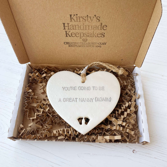 Pearlised white clay hanging heart with baby feet cut out of the bottom and grey personalisation, the heart is personalised with YOU’RE GOING TO BE GREAT NANNY (AGAIN)