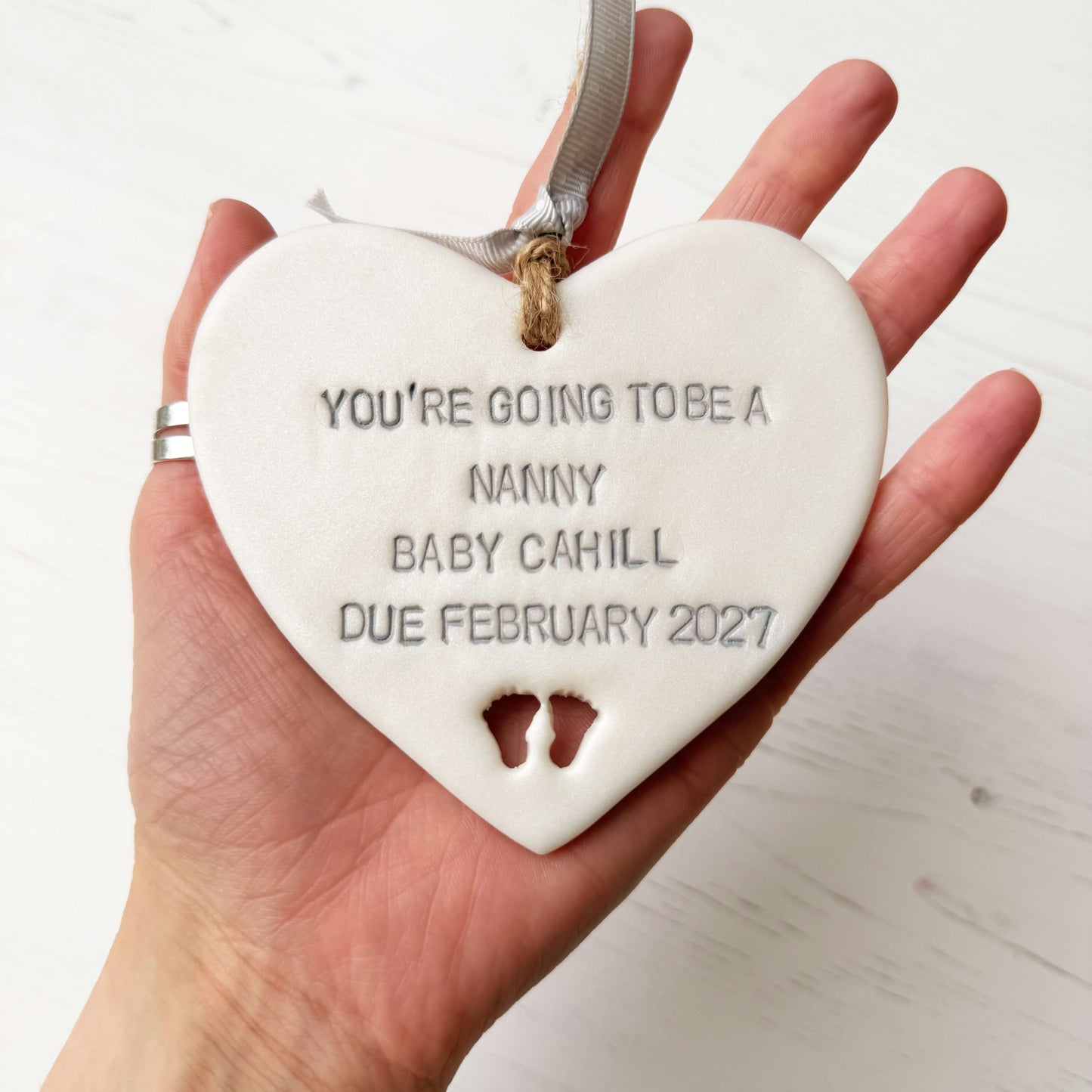 Pearlised white clay hanging heart with baby feet cut out of the bottom and grey personalisation, the heart is personalised with YOU’RE GOING TO BE A NANNY BABY CAHILL DUE FEBRUARY 2027
