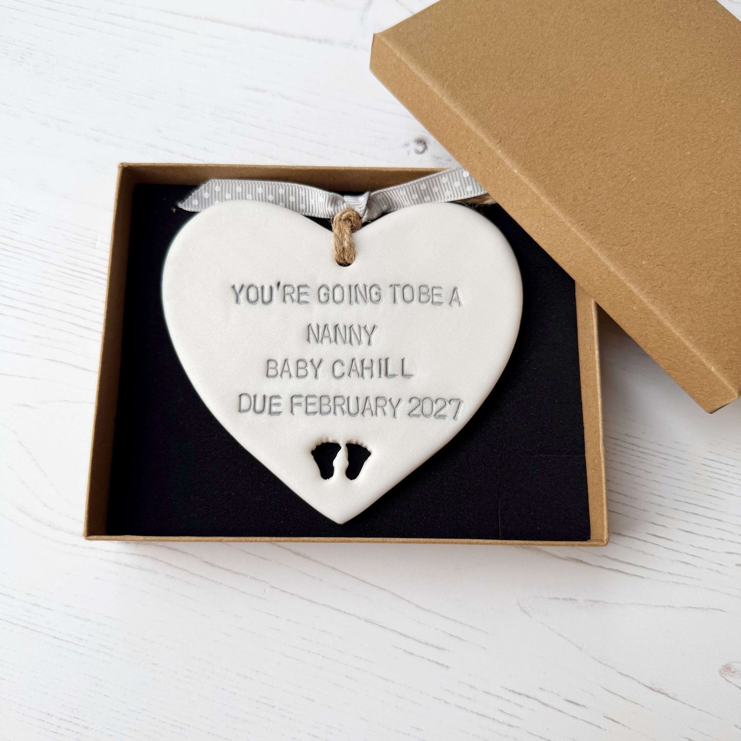 Pearlised white clay hanging heart with baby feet cut out of the bottom and grey personalisation, the heart is personalised with YOU’RE GOING TO BE A NANNY BABY CAHILL DUE FEBRUARY 2027