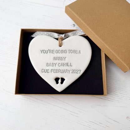 Pearlised white clay hanging heart with baby feet cut out of the bottom and grey personalisation, the heart is personalised with YOU’RE GOING TO BE A NANNY BABY CAHILL DUE FEBRUARY 2027
