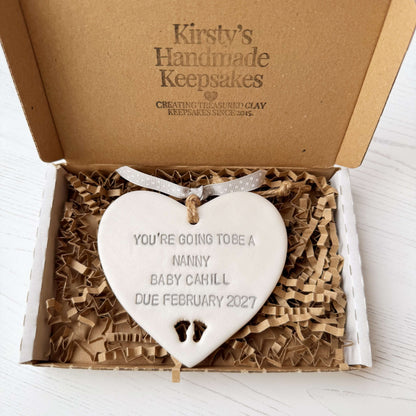 Pearlised white clay hanging heart with baby feet cut out of the bottom and grey personalisation, the heart is personalised with YOU’RE GOING TO BE A NANNY BABY CAHILL DUE FEBRUARY 2027