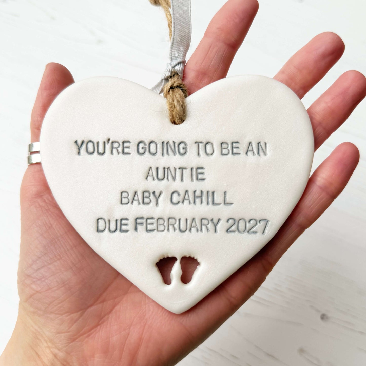 Pearlised white clay hanging heart with baby feet cut out of the bottom and grey personalisation, the heart is personalised with YOU’RE GOING TO BE AN AUNTIE BABY CAHILL DUE FEBRUARY 2027