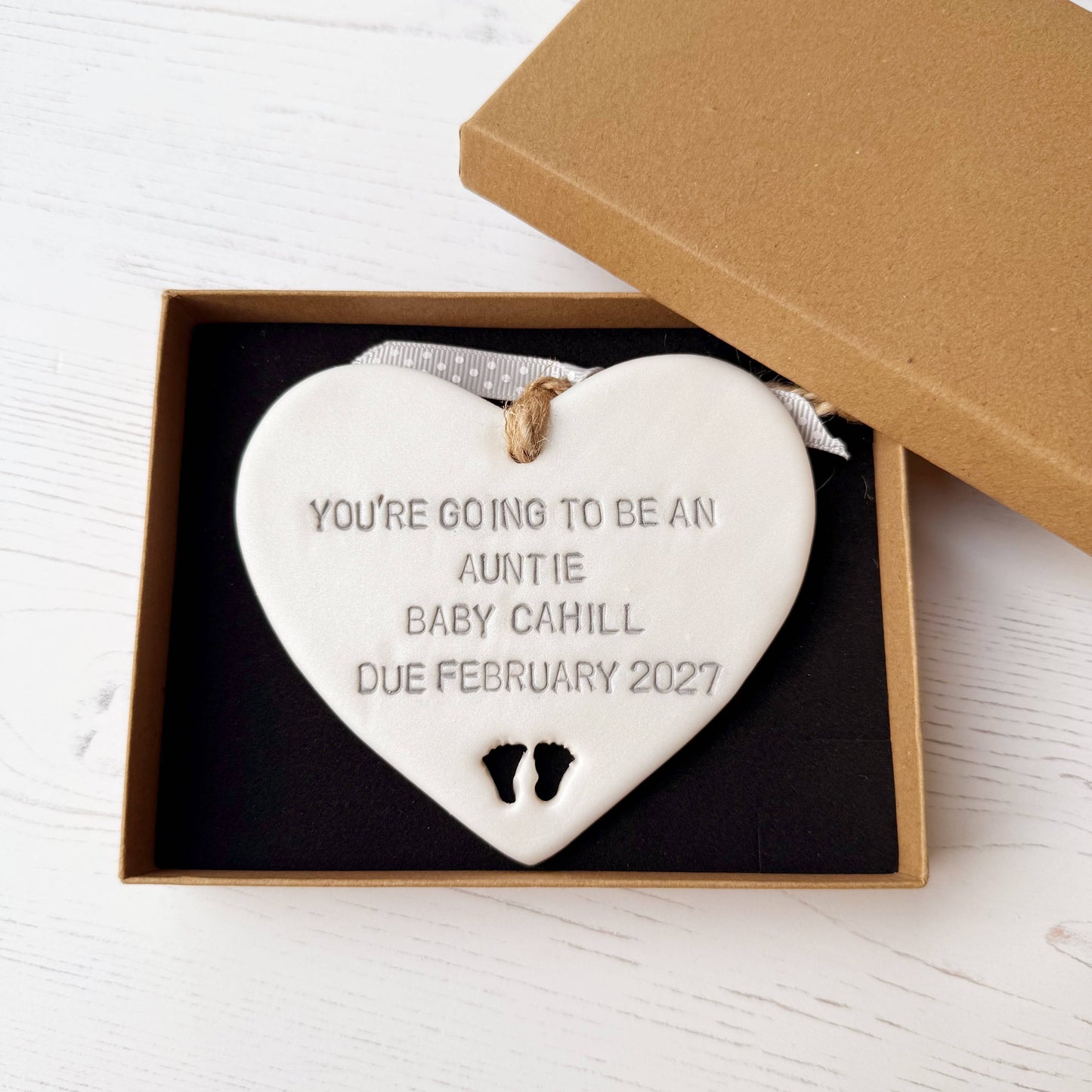 Pearlised white clay hanging heart with baby feet cut out of the bottom and grey personalisation, the heart is personalised with YOU’RE GOING TO BE AN AUNTIE BABY CAHILL DUE FEBRUARY 2027