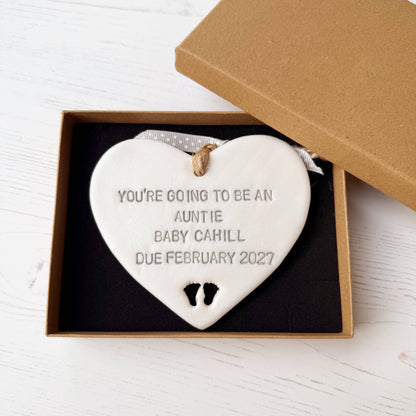 Pearlised white clay hanging heart with baby feet cut out of the bottom and grey personalisation, the heart is personalised with YOU’RE GOING TO BE AN AUNTIE BABY CAHILL DUE FEBRUARY 2027