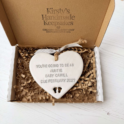 Pearlised white clay hanging heart with baby feet cut out of the bottom and grey personalisation, the heart is personalised with YOU’RE GOING TO BE AN AUNTIE BABY CAHILL DUE FEBRUARY 2027