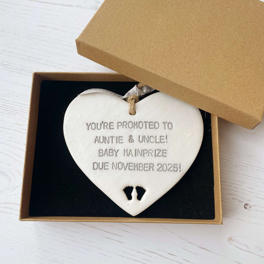 Pearlised white clay hanging heart with baby feet cut out of the bottom and grey personalisation, the heart is personalised with YOU’RE PROMOTED TO AUNTIE & UNCLE! BABY MAINPRIZE DUE NOVEMBER 2025!