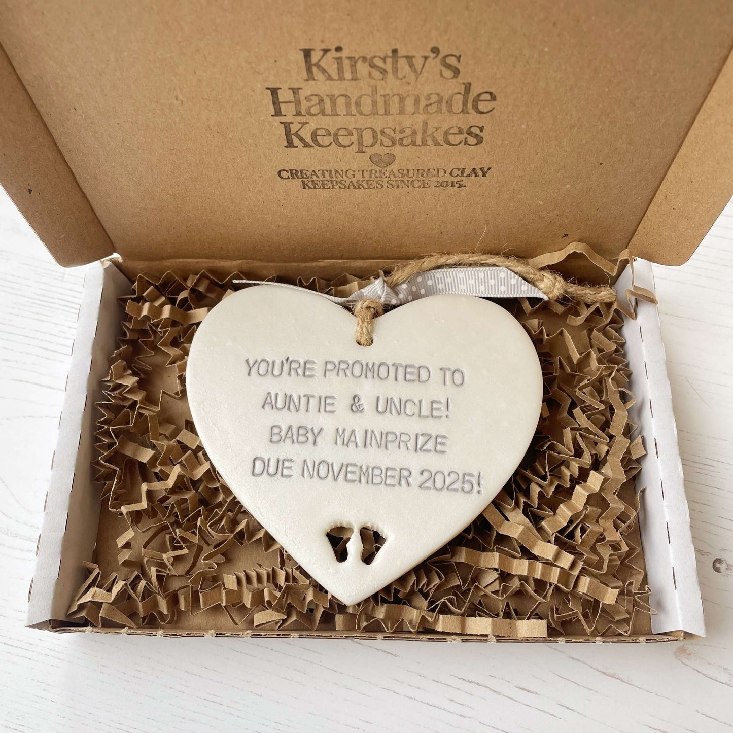 Pearlised white clay hanging heart with baby feet cut out of the bottom and grey personalisation, the heart is personalised with YOU’RE PROMOTED TO AUNTIE & UNCLE! BABY MAINPRIZE DUE NOVEMBER 2025!