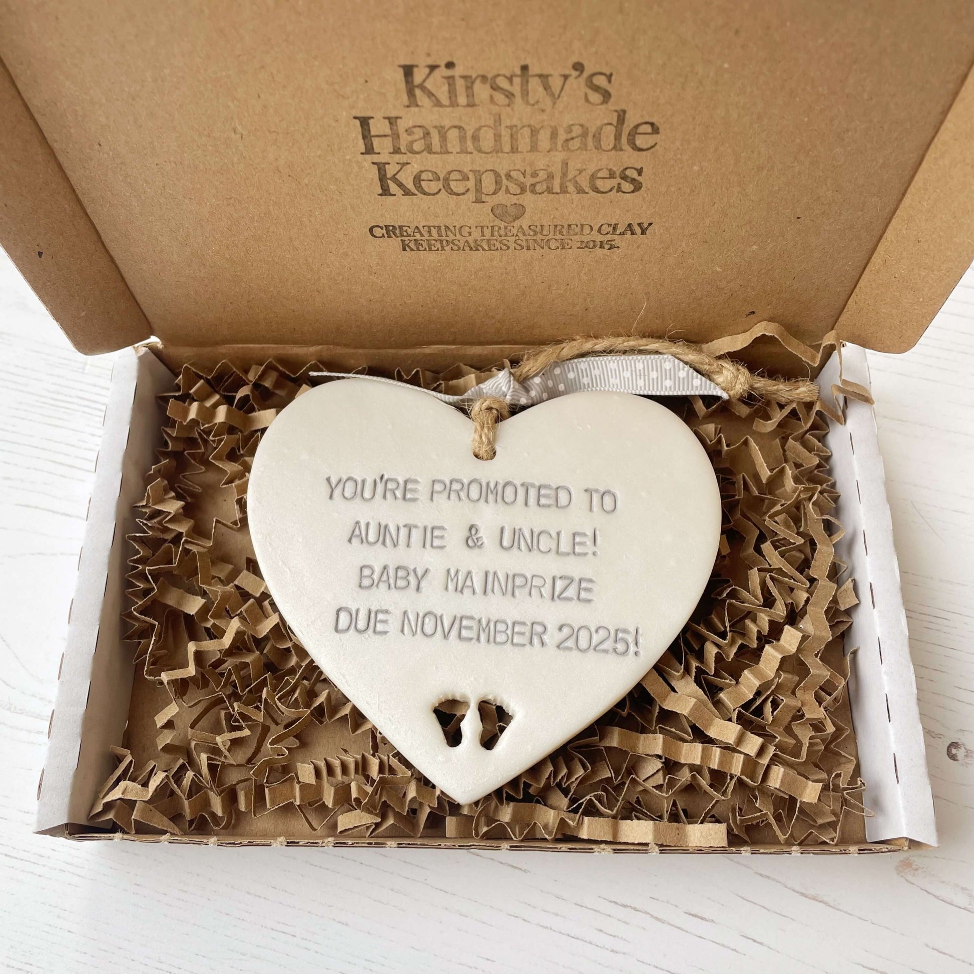 Pearlised white clay hanging heart with baby feet cut out of the bottom and grey personalisation, the heart is personalised with YOU’RE PROMOTED TO AUNTIE & UNCLE! BABY MAINPRIZE DUE NOVEMBER 2025!