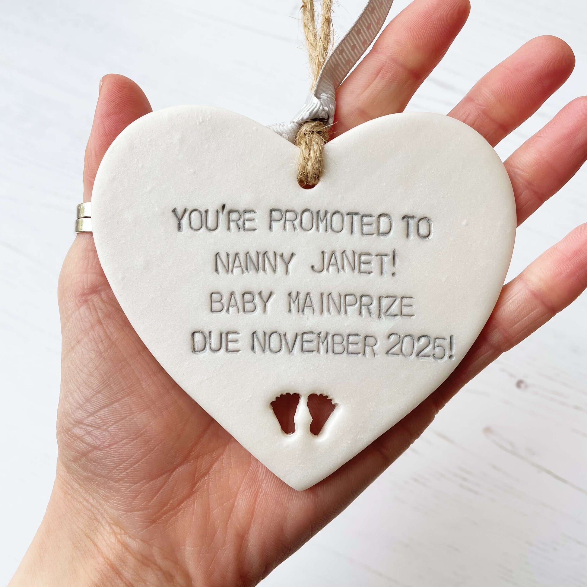 Pearlised white clay hanging heart with baby feet cut out of the bottom and grey personalisation, the heart is personalised with YOU’RE PROMOTED TO NANNY JANET! BABY MAINPRIZE DUE NOVEMBER 2025!
