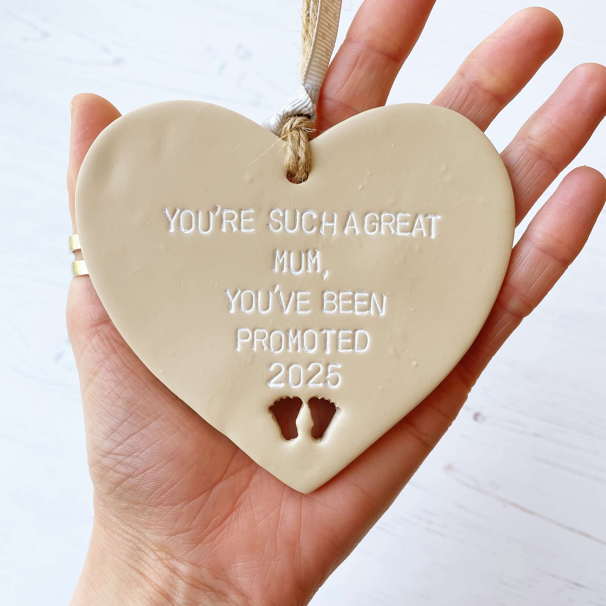 Beige clay hanging heart with baby feet cut out of the bottom and white personalisation, the heart is personalised with YOU’RE SUCH A GREAT MUM, YOU’VE BEEN PROMOTED 2025