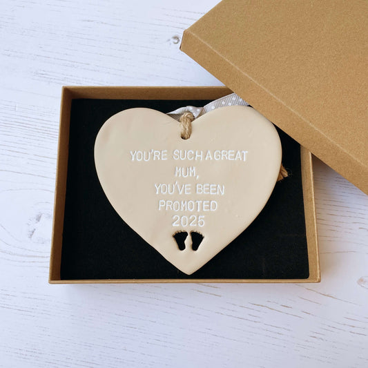 Beige clay hanging heart with baby feet cut out of the bottom and white personalisation, the heart is personalised with YOU’RE SUCH A GREAT MUM, YOU’VE BEEN PROMOTED 2025