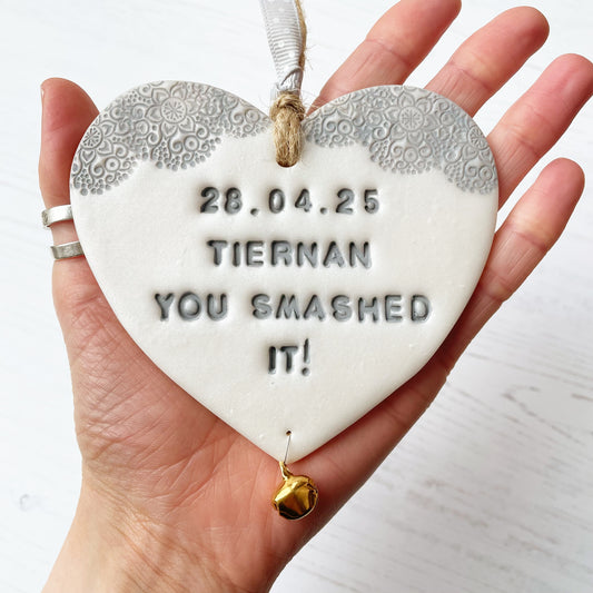 Personalised cancer survivor gift, pearlised white clay hanging heart with a grey lace edge at the top of the heart and a gold bell hanging below, the heart is personalised with 28.04.25 TIERNAN YOU SMASHED IT!