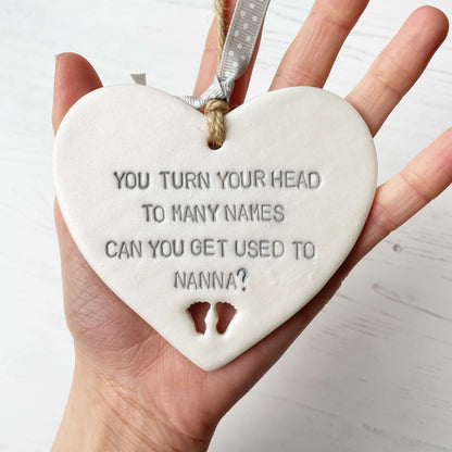 Pearlised white clay hanging heart pregnancy reveal with baby feet cut out of the bottom and grey personalisation, the heart is personalised with YOU TURN YOUR HEAD TO MANY NAMES CAN YOU GET USED TO NANNA?