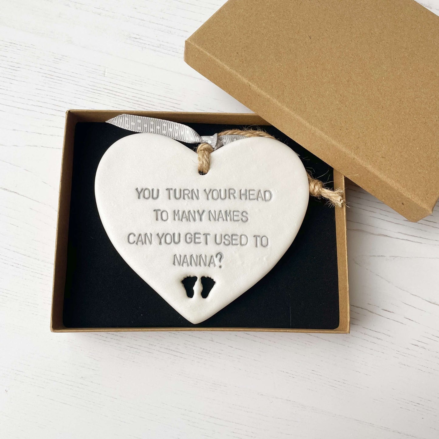Pearlised white clay hanging heart pregnancy reveal with baby feet cut out of the bottom and grey personalisation, the heart is personalised with YOU TURN YOUR HEAD TO MANY NAMES CAN YOU GET USED TO NANNA?