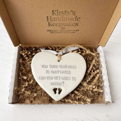 Pearlised white clay hanging heart pregnancy reveal with baby feet cut out of the bottom and grey personalisation, the heart is personalised with YOU TURN YOUR HEAD TO MANY NAMES CAN YOU GET USED TO NANNA?
