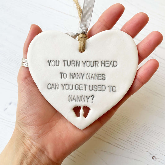 Pearlised white clay hanging heart pregnancy reveal with baby feet cut out of the bottom and grey personalisation, the heart is personalised with YOU TURN YOUR HEAD TO MANY NAMES CAN YOU GET USED TO NANNY?