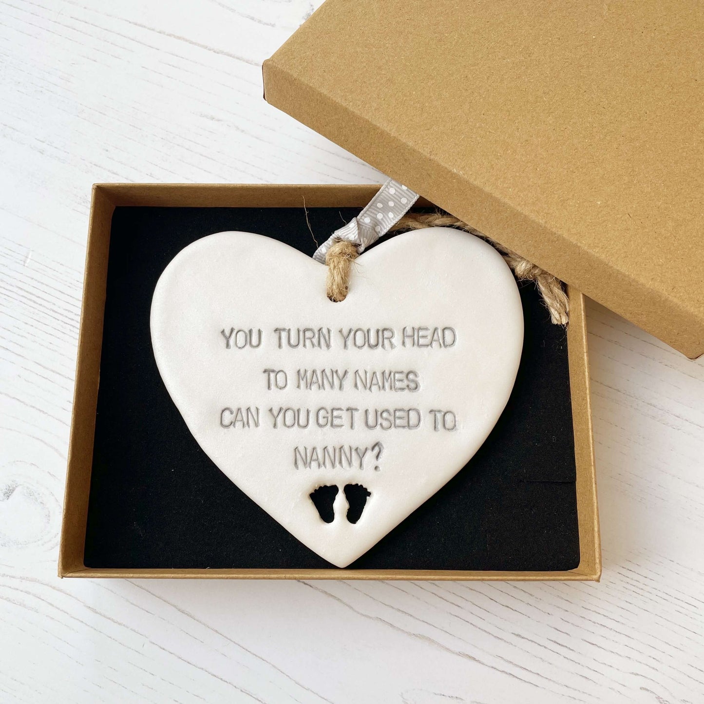 Pearlised white clay hanging heart pregnancy reveal with baby feet cut out of the bottom and grey personalisation, the heart is personalised with YOU TURN YOUR HEAD TO MANY NAMES CAN YOU GET USED TO NANNY?