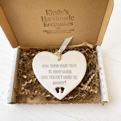 Pearlised white clay hanging heart pregnancy reveal with baby feet cut out of the bottom and grey personalisation, the heart is personalised with YOU TURN YOUR HEAD TO MANY NAMES CAN YOU GET USED TO NANNY?