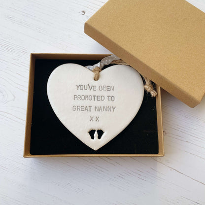 Pearlised white clay hanging heart with baby feet cut out of the bottom and grey personalisation, the heart is personalised with YOU’VE BEEN PROMOTED TO GREAT NANNY XX
