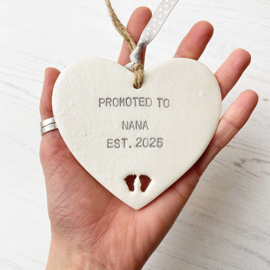 Pearlised white clay hanging heart with baby feet cut out of the bottom and grey personalisation, the heart is personalised with PROMOTED TO NANA EST. 2025