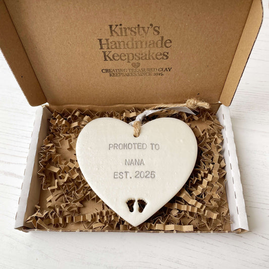 Pearlised white clay hanging heart with baby feet cut out of the bottom and grey personalisation, the heart is personalised with PROMOTED TO NANA EST. 2025